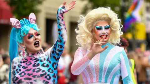 Pacemaker Two people dressed in bright colours on a float at a festival. One is wearing a blue wig and wearing pink and blue cheetah print and big silver and pink earrings. The other is wearing a blond wig and wearing pink, blue and silver. Both are wearing make-up.