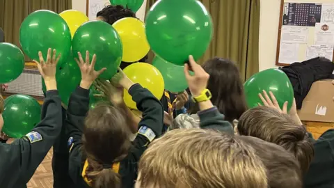 London Ambulance Service Children in green Scout uniforms have their arms in the air and are holding up green and yellow balloons