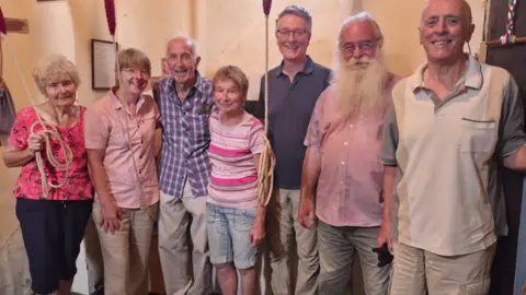 Seven men and women, all middle-aged or older, in a church tower, some holding bell ropes, and smiling to camera. 