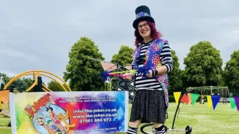 Daventry Town Council Performer with long purple hair wearing a black top hat and glasses, a dark skirt and black and white top with a multicoloured waistcoat. The performer is holding some coloured plastic straws and is standing next to a banner advertising "the Joker". There is a unicycle behind the performer, some coloured bunting and trees in the background.