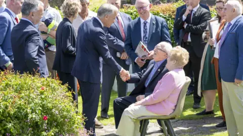 PA Media Canadian Prime Minister Mark Carney is shaking hands with two pensioners. The couple are sitting on chairs outside while Carney who is standing up is leaning down to greet them. They are outside and surrounded by a large group of people. 