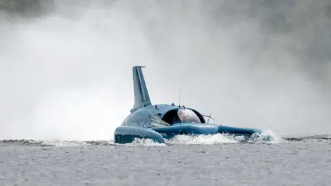 Getty Images Close-up of Bluebird operating on Loch Fad. It is a craft that looks like a cross between a plane and a boat. It is painted blue. Spray is shooting up behind it.