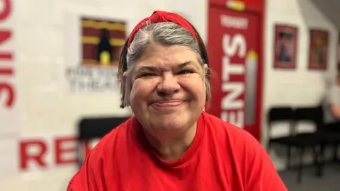 A women with short grey hair looking and smiling at the camera. she has a red band in her hair and a red t-shirt on. 