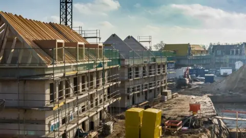A stock image showing a new housing development being built, with scaffolding surrounding the buildings and stacked piles of building materials on muddy land.