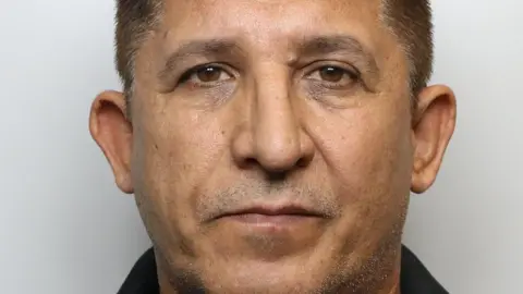 A mugshot of Mohammed, who has short, dark hair that is neatly trimmed and evenly cut across the top and sides. His face is fully visible, with symmetrical features, dark eyebrows, and eyes looking directly toward the camera. His facial hair is minimal, with slight stubble visible around the chin and jawline area.