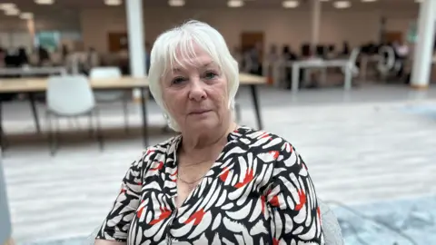 Kate Bradbrook/BBC A woman in a white, red and black dress sits in an office looking at the camera. 