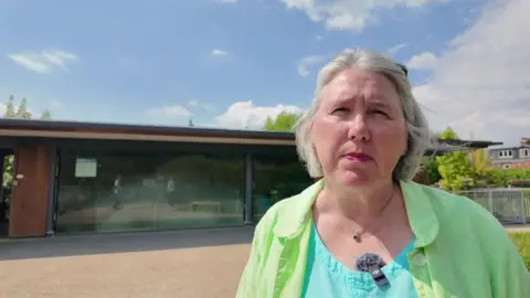 Barbara Hammond has medium length grey hair and is wearing a green shirt over a blue t-shirt. She is standing in front of Osney Lock Hydro.