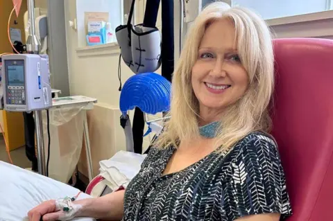 Helene Rose A woman with long blonde hair and a black short-sleeved top sits in a hospital chair, hooked up to a machine. She is smiling at the camera.