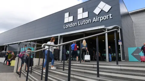 LLA The outside of London Luton Airport, with steps. a large LLA sign, with people walking into the building.