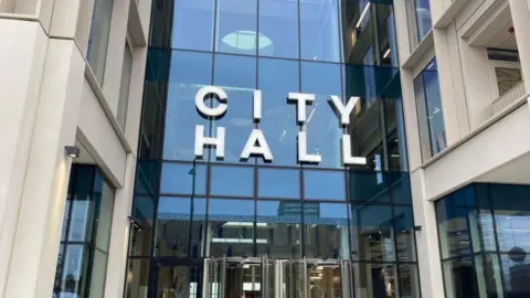 The image shows the entrance to Sunderland City Council. It's a modern building with the words City Hall above a glass fronted entrance and revolving doors 