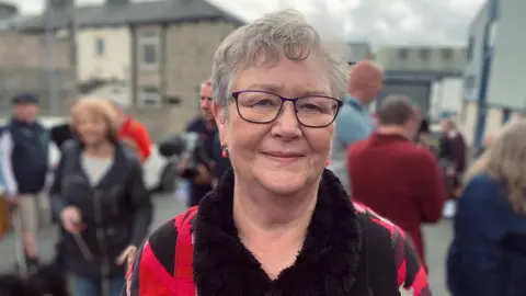 BBC/Phil Cunliffe Ruth Angus wears a red and black jumper and has short grey hair with dark-rimmed glasses