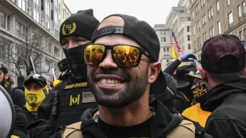 Getty Images Former Proud Boys leader Enrique Tarrio is shown smiling while wearing mirrored sunglasses and back and yellow protective clothing. He is standing outside Harry's bar in Washington, DC during a protest on 12 December, 2020