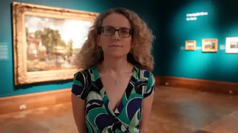 Qays Najm/BBC A woman wearing glasses stands in the middle of an art exhibition to pose and smile at the camera. She has long curly blonde hair and is wearing a green and blue dress.
