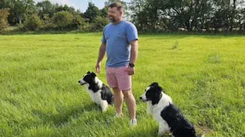 Ben Smith Ben is wearing a blue t-shirt and blue shorts and is a standing in a field of long green grass with his two dogs sitting either side of him.