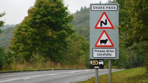 A sign for Snake Pass with a wiggly road warning and a red triangle showing livestock, and a please drive carefully plea, next to a section of road with looming Peak District landscape in the background