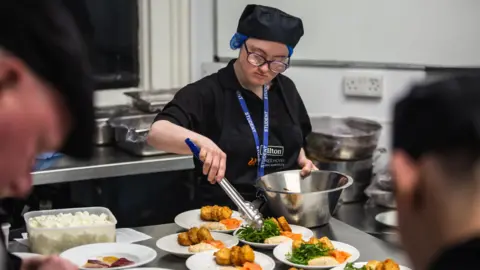 Andy Barnham Students working in a hotel kitchen. Two students in the foreground are blurred. The student in the background is using serving tongs to put salad on a plate of food. There are several plates on the table. The student is wearing a black t-shirt, black hat and glasses.