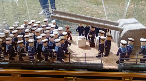 The panel knitter by Rosemary Fielder and her friends inside a glass case. A large number of sailors in uniform stands on one side, in front of a tank. A press photographer stands in front of them, taking a picture of Churchill standing on a pulpit, surrounded by other officials.