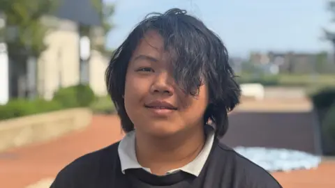 Bofeng is smiling at the camera. He has black wavy hair with his fringe covering one eye. He is wearing a white shirt with a dark sweater and is standing in the college grounds.