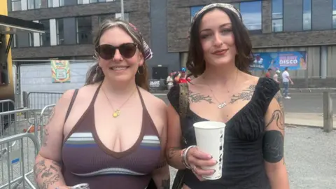 Two women smile into the camera. The woman on the left wears large sunglasses, a banana and a gold locket necklace with a brown vest top. The woman on the right has black hair, is also wearing a bandana and has a silver necklace which says M on it and is wearing a black top and holding a large white cup. Metal railings and a street can be seen behind them.