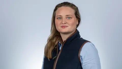 Amy Morris Amy Morris, who has shoulder length brown hair, poses for the camera. She is wearing a light blue shirt and a dark blue gilet
