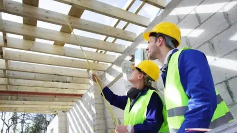 A woman in a yellow hard hat and green high-vis jacket stands next to a man in the same gear. Both are looking up, away from the camera at the wooden slats of a roof being built on top of grey-coloured bricks. The woman holds a tape measure with one end of it leaning against one of the slats