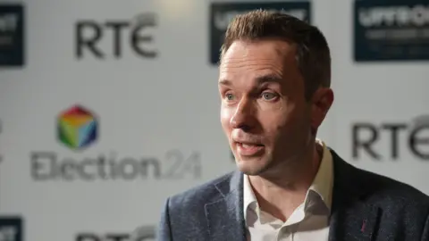 PA Cian O'Callaghan with short brown hair and blue eyes speaking to the camera. He is wearing a dark blazer and a white shirt.