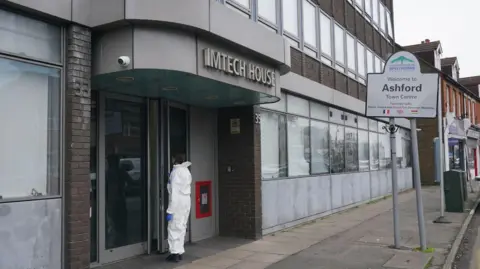 A large building in a high street with a person in a white forensic suit entering the main front door