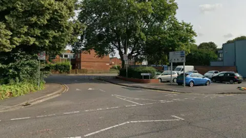 Street view of the junction with cars seen in a car park and trees on either side of the road