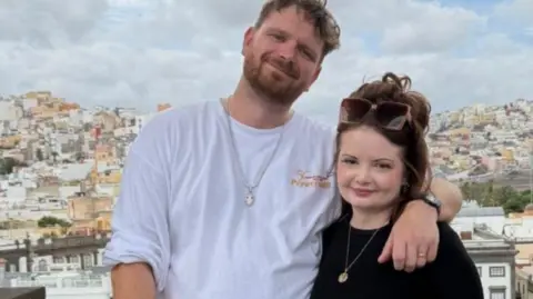 Cancer Research UK handout Jak with brown wavy hair and beard wearing a white t-shirt has his arm around his wife, Gemma Coleman, who has long brown hair with sunglasses resting on her head and wearing a black top. They are on holiday abroad with houses and a bridge in the background.