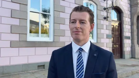 Deputy Max Andrews is wearing a dark blue blazer, white shit and a tie with blue and white stripes.