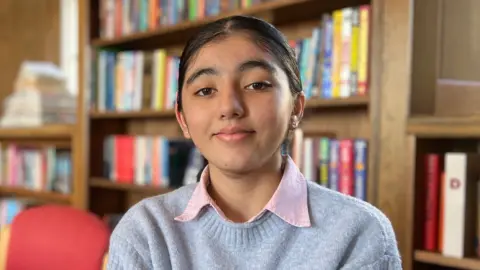 Sofiya sits in a library with bookshelves filled with colourful books in the background. She is wearing a light grey Tommy Hilfiger sweatshirt with a large red, white, and blue logo on the front and a collared shirt underneath. Wooden bookshelves and red chairs surround the seating area.