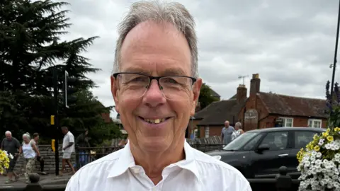 George Cowcher has grey hair and black glasses and is wearing a white shirt. In the background is a car passing by as well as pedestrians walking alongside a stone wall.