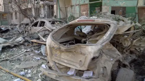 A landscape image of rubble on the street and two cars can be seen heavily damaged.