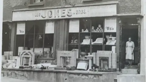 Geo Jones & Bros A black-and-white photo taken approximately 100 years ago: a woman stands at the doorway of a shop selling fireplaces. The name Geo Jones & Bros is written above the shop window.. A large assortment of fireplaces and basins can be seen both outside and inside the shop. 