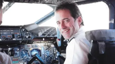 An old photograph of John Tye in the cockpit of a Concorde plane. He is sitting in the pilot's seat, with the steering wheel in front of him, dressed in a white shirt. He is looking over his shoulder and smiling at the camera. 
