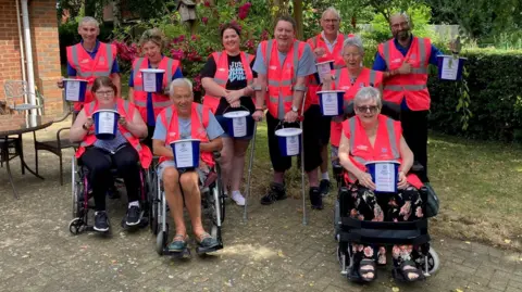 Bedford MS Therapy Centre A group of people, in pink high-vis jackets, all looking at the camera and smiling. They are all holding blue buckets with writing on. Three are in wheelchairs and one person has crutches. They are by a brick building and a bush, with trees and foliage behind them. 