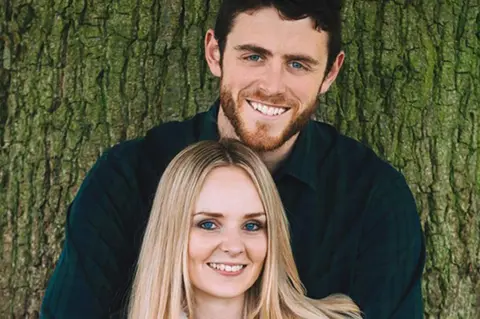 PA Andrew holds Lissie from behind as they pose for a happy portrait in front of a tree.