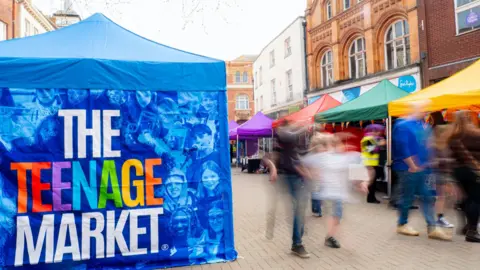 A blue gazebo on the left of the image which has the faces of young people on it and the words The Teenage Market. The people are blurred on a long focus. on the right of the image are yellow, green, red and purple gazebos with market stalls.