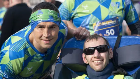PA Media Kevin Sinfield (left) and Rob Burrow (right) wearing rugby kit. Sinfield is leaning towards Burrow's adjustable chair. Both men are smiling at the camera.