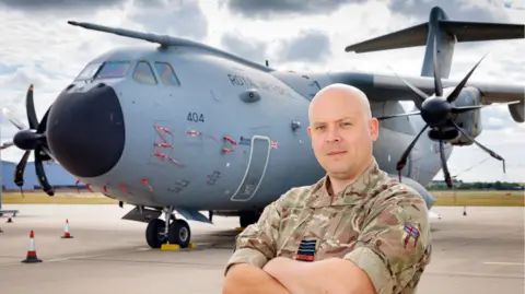 Cameron Thomas MP in military fatigues in the foreground with his arms crossed. He is stood in front of a grey Hercules plane.