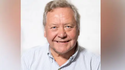A man in a striped blue-and-white shirt with an open collar smiles against a white backdrop. He has grey hair and blue eyes.
