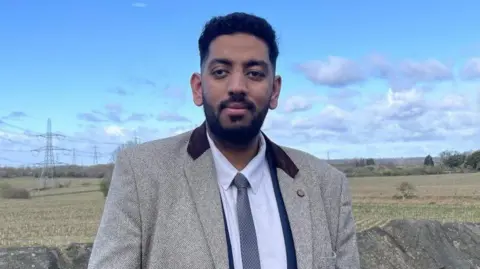A man, wearing a light grey blazer with dark coloured collar over a white shirt and blue tie, looks at the camera. There is a stone wall behind him, as well as a field with trees and bushes.