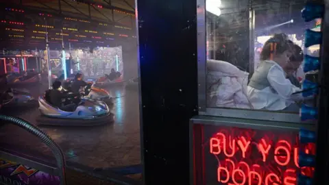 A child sits with an adult in a ticket box on a fairground, with people enjoying the dodgems to the left of the photo.