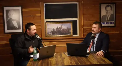 Contra Mundum Dua pria duduk di depan mikrofon dan komputer, dengan karya seni patriotik di belakangnya, termasuk salinan lukisan terkenal George Washington selama Perang Revolusi AS dan mantan presiden Richard Nixon dan James Polk