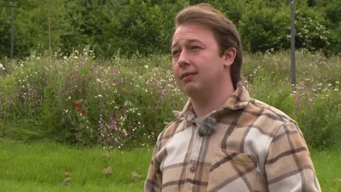 Jordan Perris, who has short brown hair and is wearing a checked brown shirt, is being interviewed. He is standing on a section of grass with flowers and trees behind him.