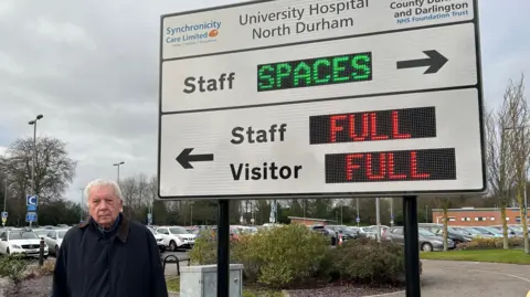 Parish councillor Grenville Holland, wearing a dark winter coat, is standing in front of a sign for the car park. A full car park is visible in the background. The sign itself if white and has digital elements. The digital parts display the words "full" in red next to an arrow to visitor and staff parking. An arrow pointing in the opposite direction labelled 'staff' says 'spaces' in green.