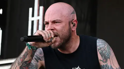 Ryan Clark, who is the lead singer of metal band Demon Hunter. He is crouched on stage wearing a black vest and camouflage trousers and is holding a microphone.