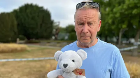 Stuart Woodward/BBC Stephen Goodsell wears a blue T-shirt while holding a grey teddy bear. He has sunglasses on top of his head and looks solemn. He is standing in front of a grassed area that has been taped off by police.