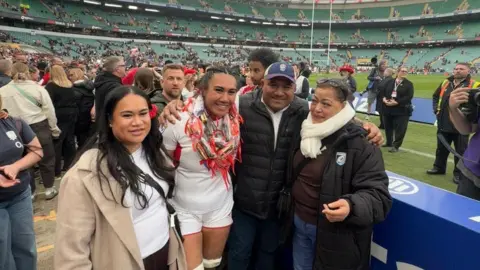 Haineala Lutui Lutui pictured with her family at Twickenham stadium. Behind them is a giant green stadium with players and fans behind them. 