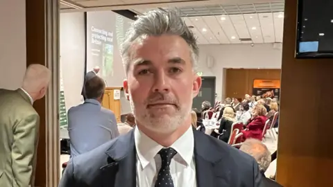 BBC/RICHARD EDWARDS An image of a man wearing a suit and tie looking straight at the camera. He has styled, grey hair and grey stubble. A busy conference hall is behind him in the background.